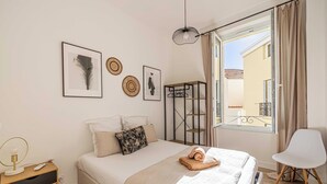 Een slaapkamer met een bed, nachtkastje, stoel, raam met gordijnen en wanddecoratie.