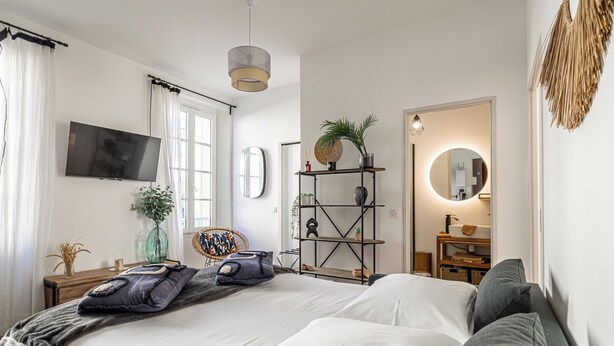 Een moderne slaapkamer met een groot bed, een aan de muur bevestigde televisie, een houten plank met planten en een spiegel met ronde verlichting.