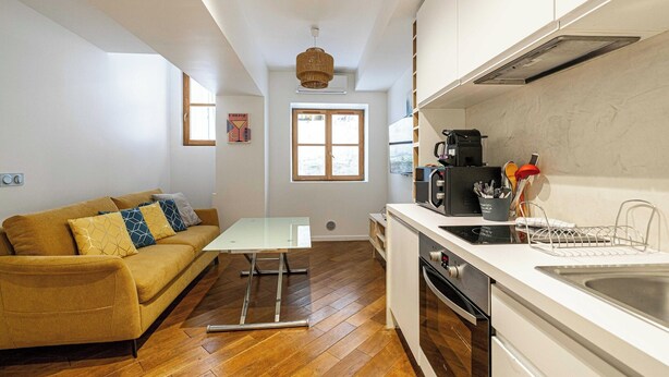 A compact kitchen with a white countertop, a built-in oven, and a coffee maker. There is a sofa with patterned cushions in the adjacent living area.