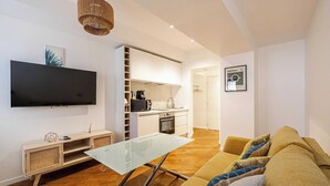 A modern living room with a flat-screen TV, a glass-top coffee table, a sofa, and a kitchen area with white cabinets and a microwave.