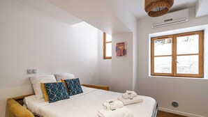 A bedroom with a bed, pillows, a bedside table, a window, and a wall-mounted air conditioner.