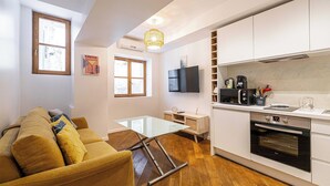 A compact living space with a sofa, a glass-top coffee table, a TV, a kitchen area with a stove, and a window.