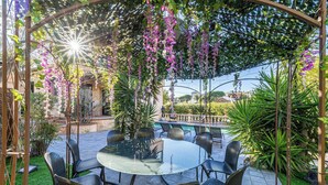 Un patio al aire libre con una mesa y sillas de vidrio redondas, rodeado de follaje exuberante y flores moradas.