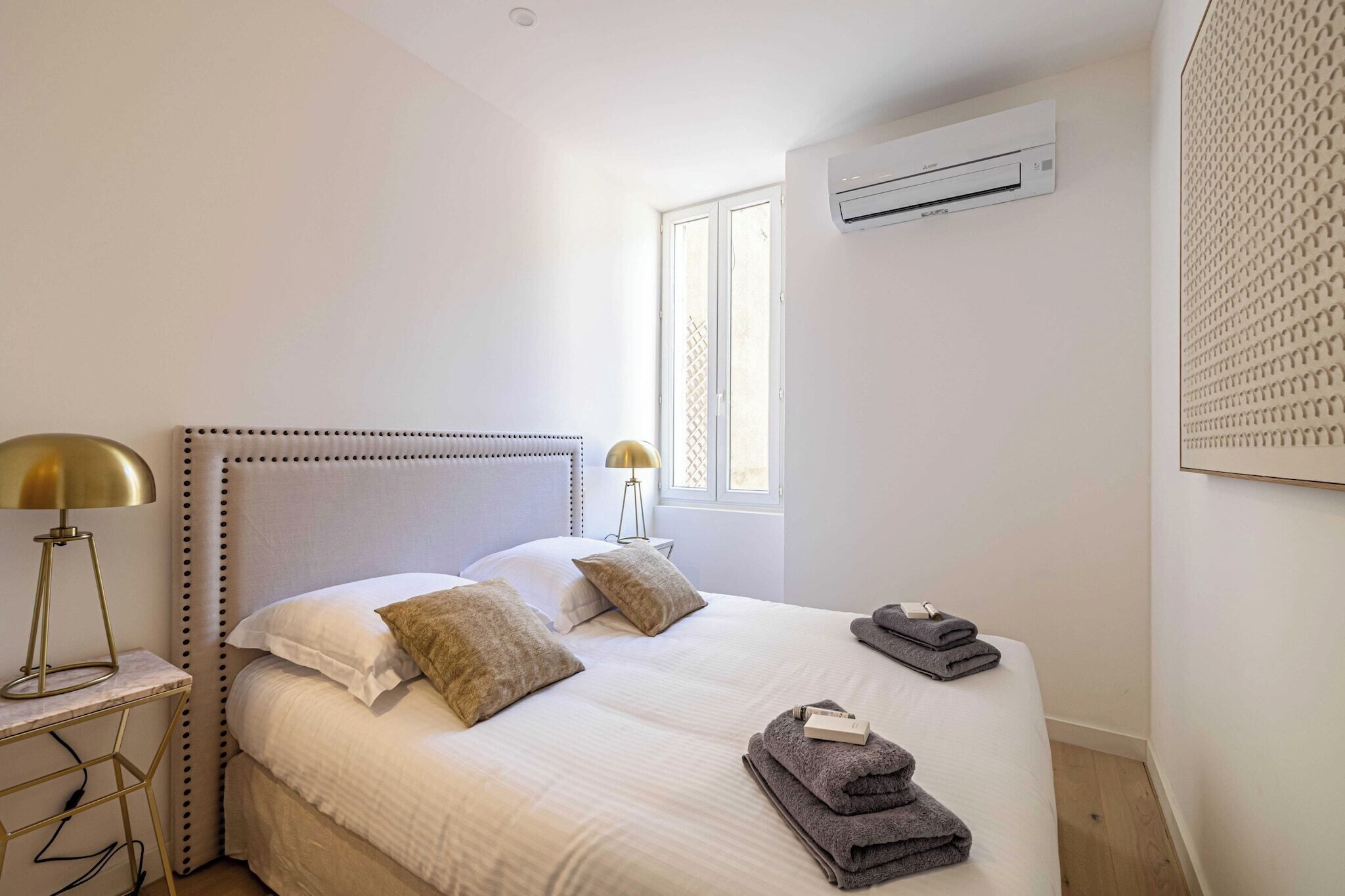 A bedroom with a bed, bedside lamp, air conditioning unit, window, and towels.