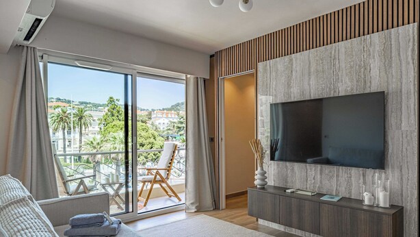 A modern hotel room with a large TV, a sofa, a balcony with a chair, and a view of the city.