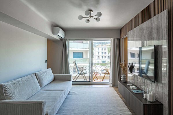 En moderne stue med sofa, tv og en balkon med udsigt til bygninger.