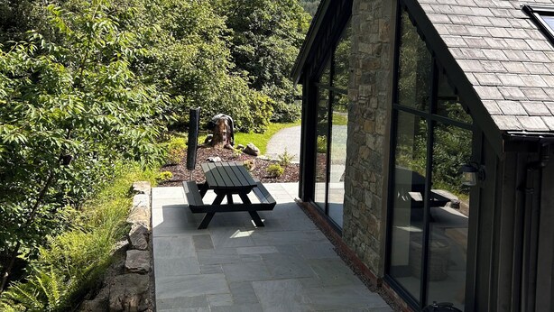 A stone building with a sloped roof and a glass door, a picnic table, and a barbecue grill outside.
