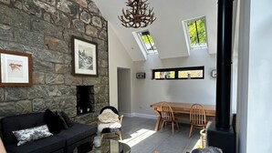 A cozy living room with a stone wall, a fireplace, a dining area with a table and chairs, and a ceiling with skylights.