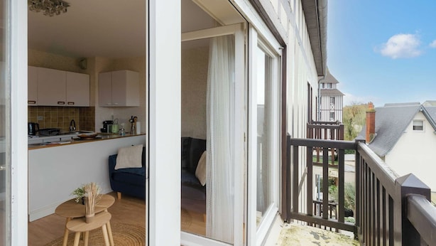 Un balcón con vistas a una zona residencial, una puerta corrediza de vidrio que da a un dormitorio y una cocina con armarios blancos y una cafetera.