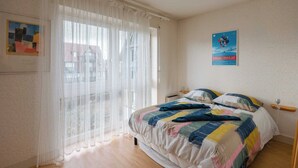 Un dormitorio con cama, ventana con cortinas translúcidas, un cartel en la pared y una mesita de noche pequeña.