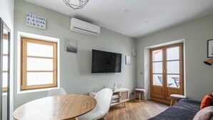 Un saló modern amb una taula de dinar de fusta, cadires blanques, un sofà negre i un televisor fixat a la paret.