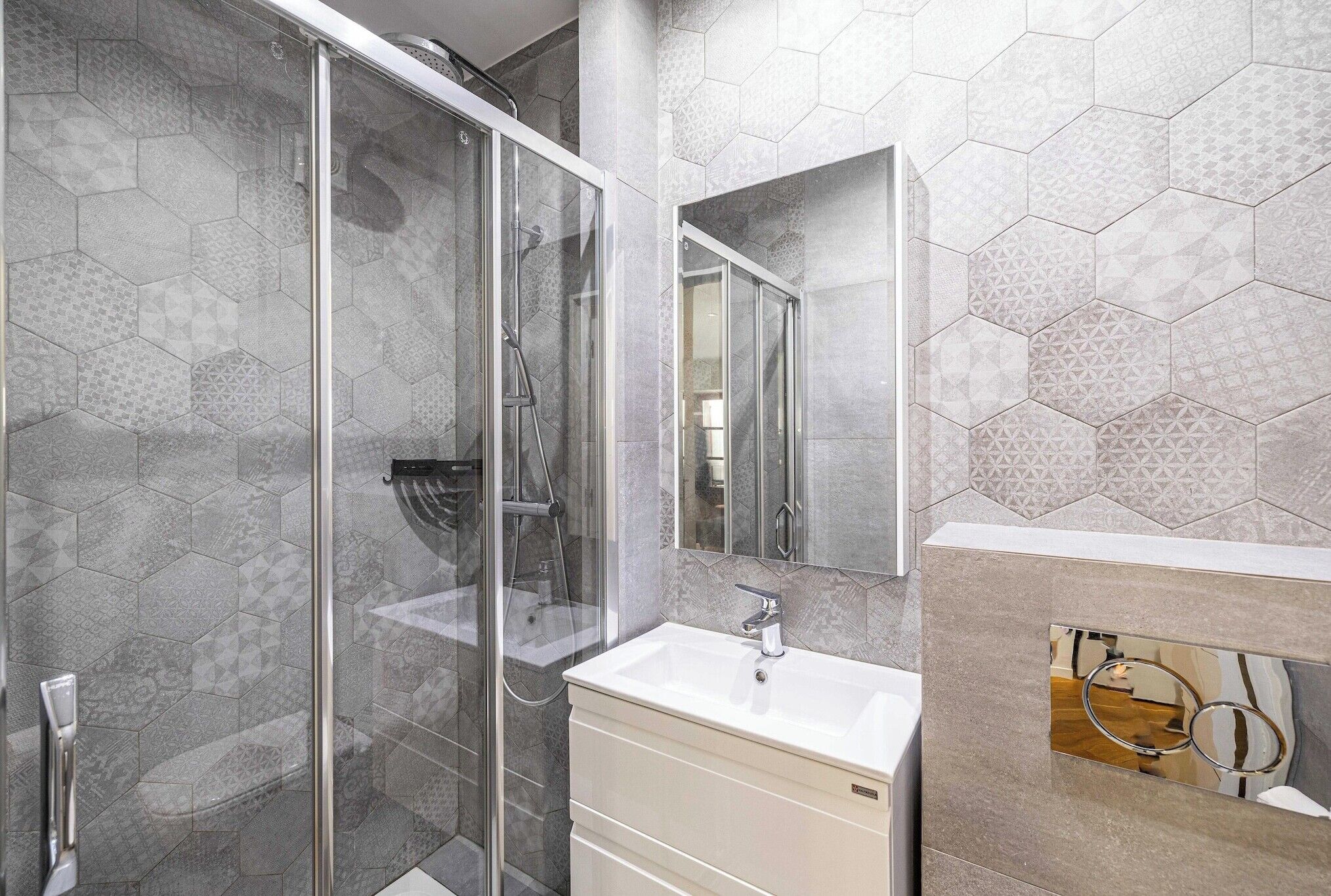 Une salle de bain moderne avec une douche à parois de verre, un lavabo blanc et un miroir agrémenté d’un pare-splash en carreaux hexagonaux.