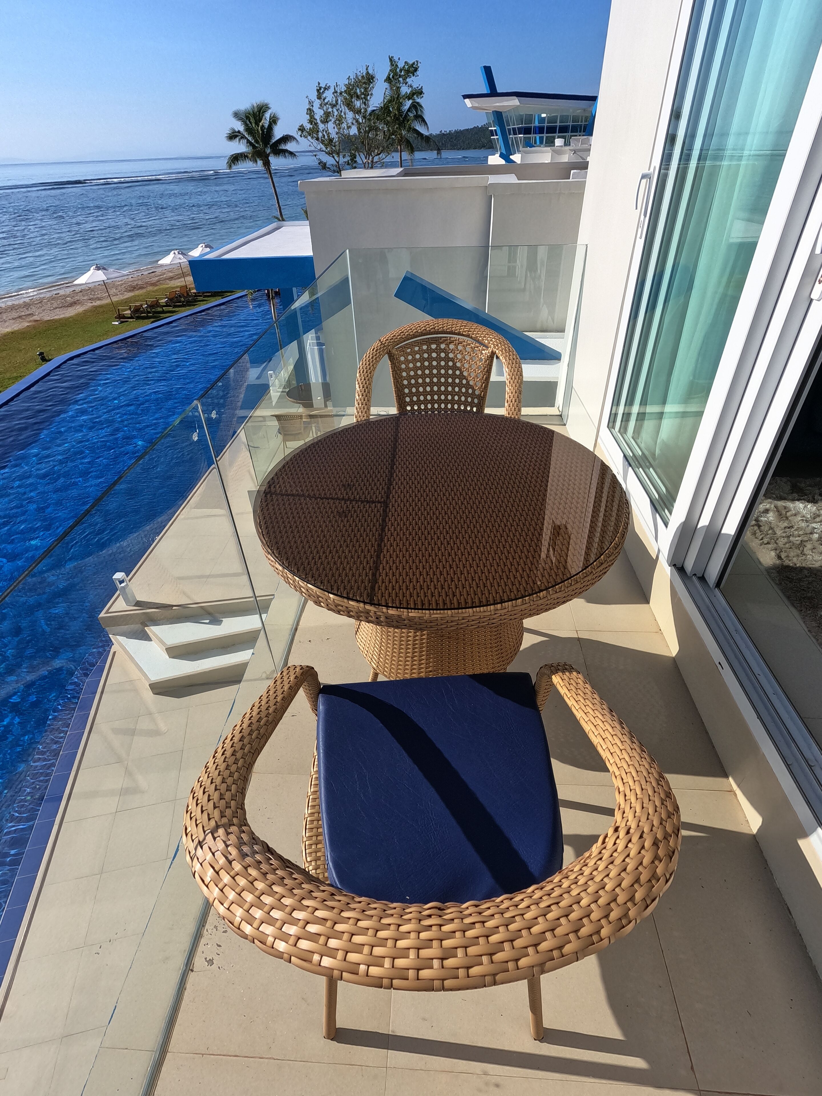 A glass-enclosed outdoor seating area with wicker furniture, overlooking a pool and ocean view.