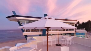 A rooftop terrace with white outdoor furniture and umbrellas, overlooking a body of water and palm trees. - Recidencia Del Hamor Beachfront (Santa Magdalena)