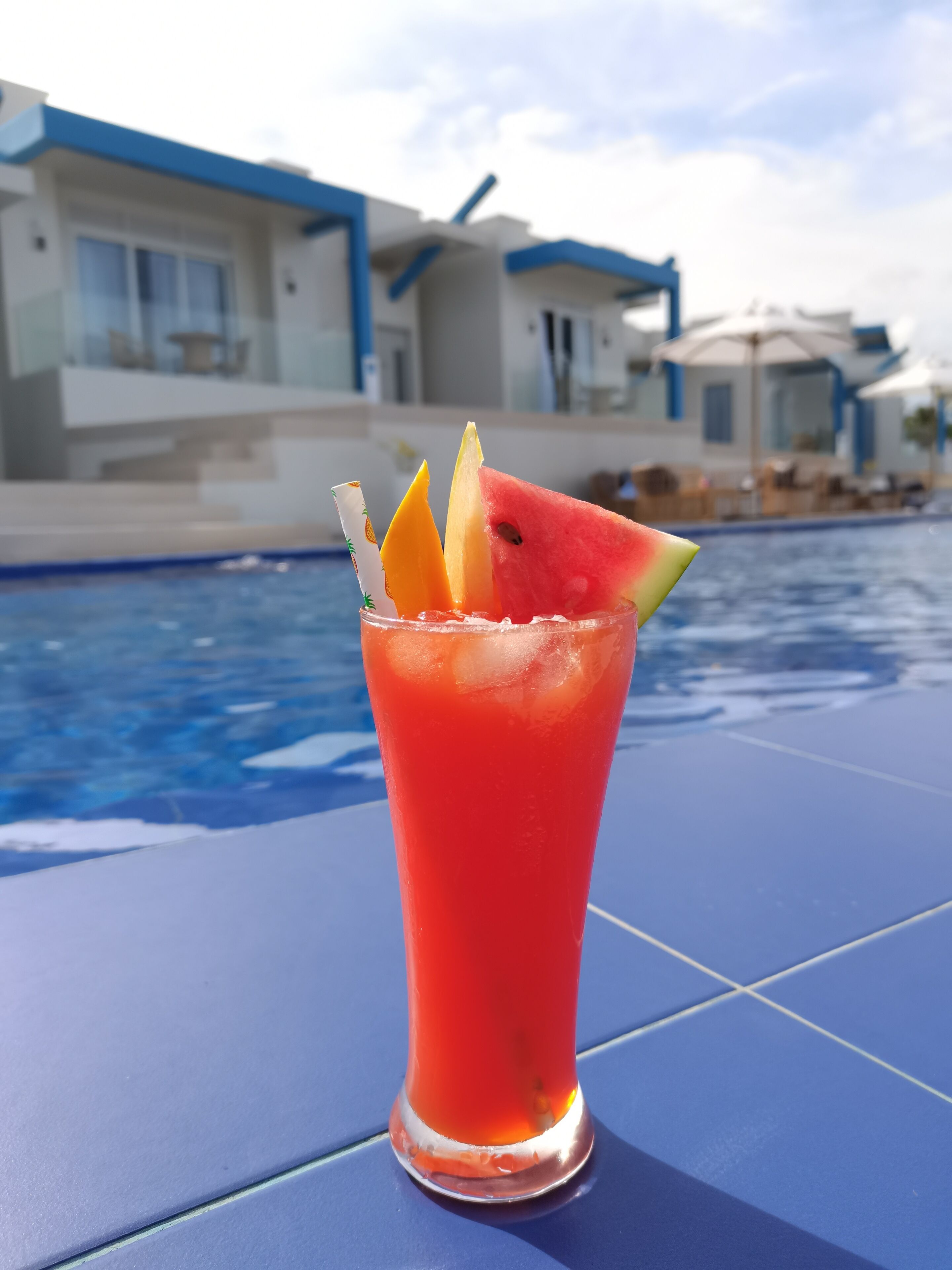Un bicchiere alto con una bevanda rossa, anguria e fettine d'arancia su un tavolo blu vicino a una piscina, con edifici moderni sullo sfondo.
