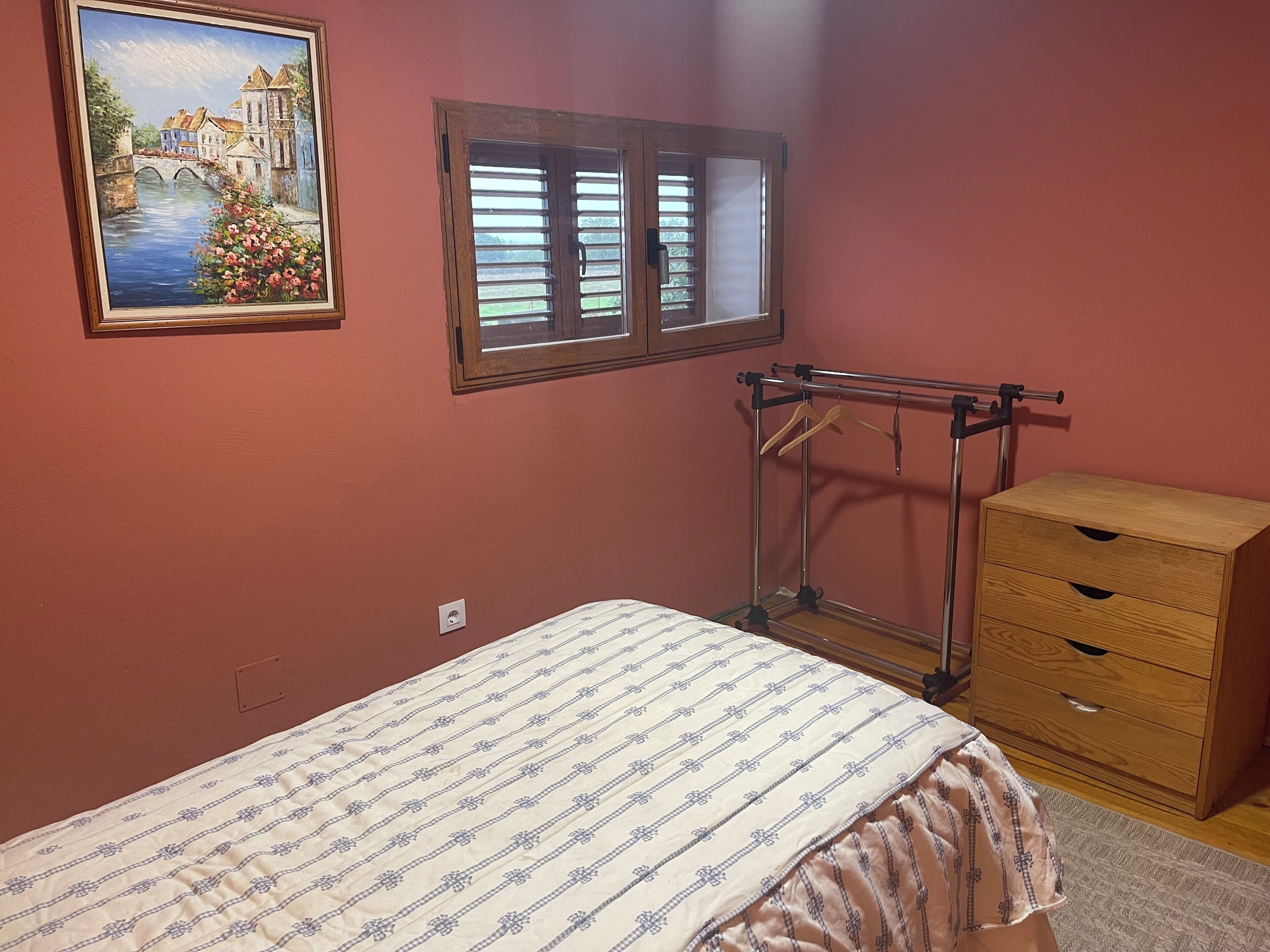 Un dormitorio con una cama, un aparador de madera, una ventana con persianas y un cuadro en la pared.