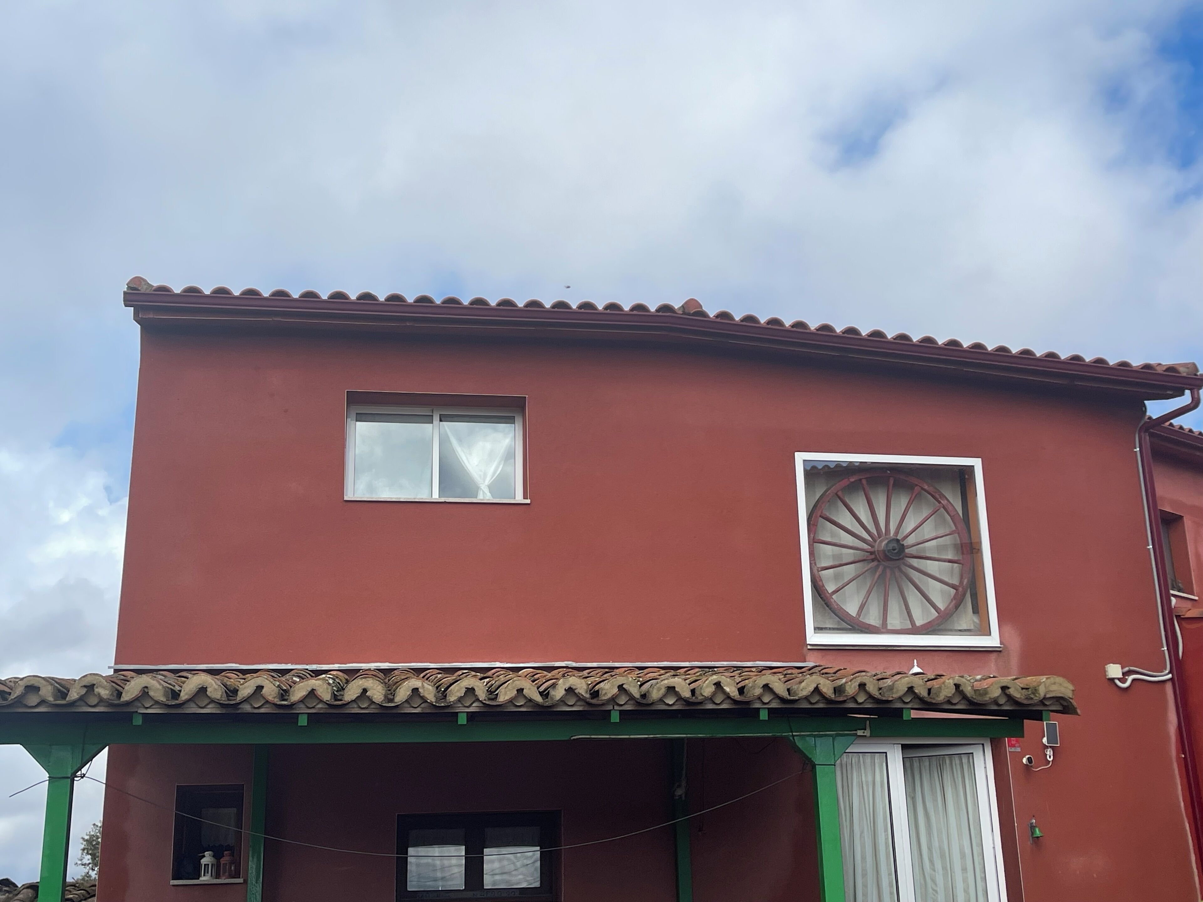 Een gebouw met twee verdiepingen, een rode gevel, een groot rond raam op de tweede verdieping en een groene veranda met een pannendak.