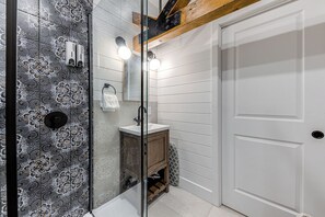 A bathroom with a patterned tile wall, a glass shower enclosure, a wooden vanity, and a white door. - Gorgeous 3 Bedroom 2-floor Old Townhouse (Québec)