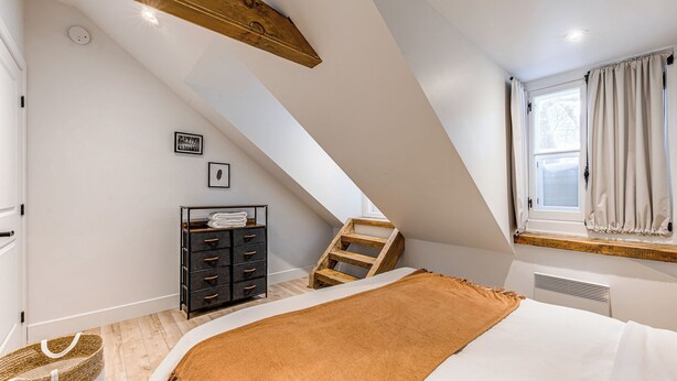 Une chambre avec un plafond en pente, un lit recouvert d’une couverture marron, une échelle en bois, une commode noire et une fenêtre avec des rideaux.