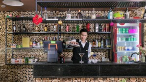A bar area with a bartender preparing a drink, shelves with various bottles and glasses, and a wall decorated with wooden logs. - SaPa Signature Inn (Sa Pa)