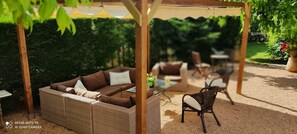 Zona d'estar exterior amb un sofà i cadires de vimetla, una taula amb un ram de flors i una estructura de pergola.