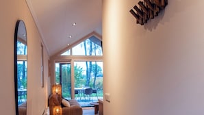 Un couloir moderne avec un plancher en bois, un grand miroir et un radiateur. On aperçoit un coin salon avec une table et des chaises à travers de grandes fenêtres.