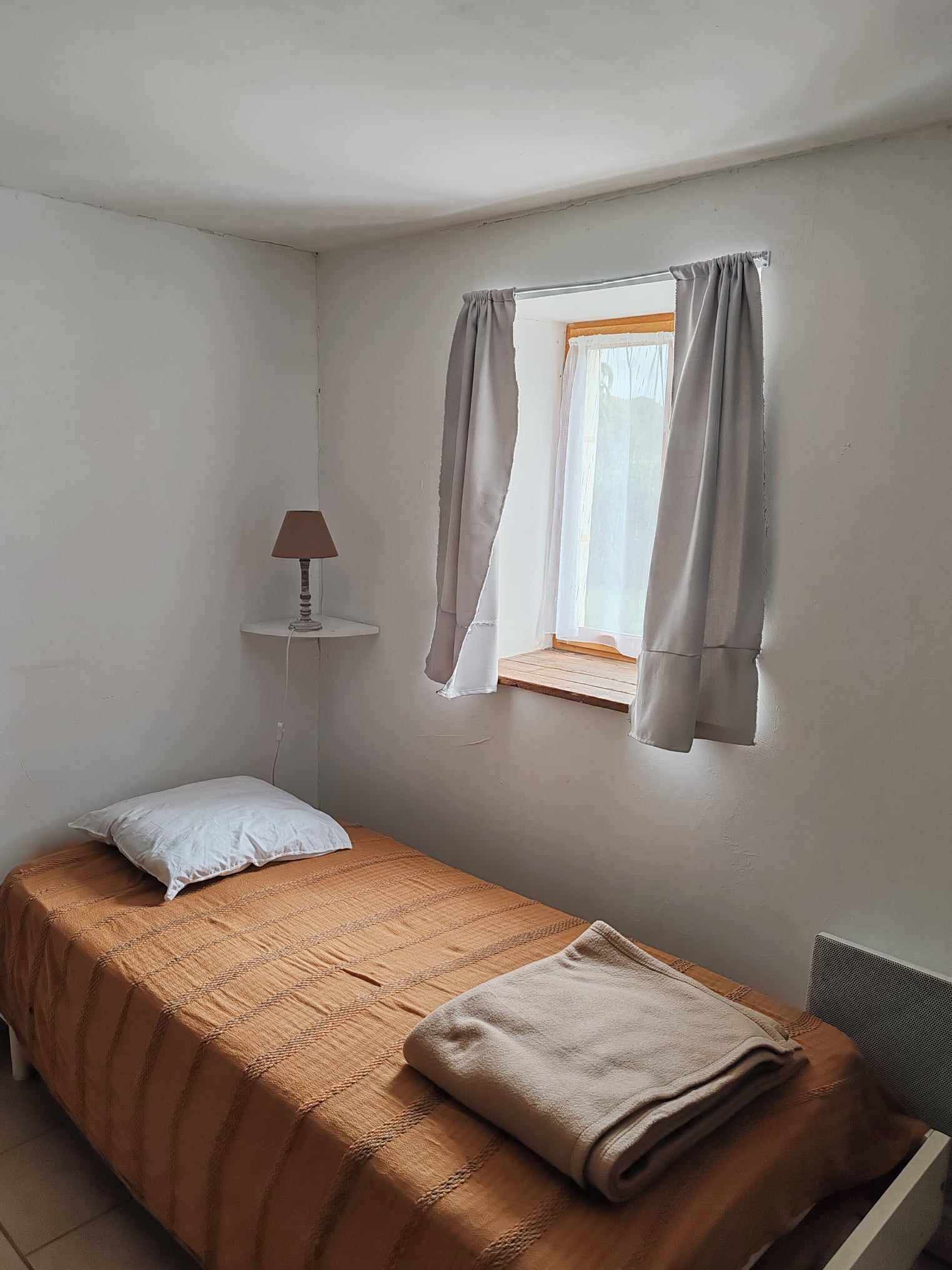 A small, single-bed room with a window, a bedside lamp, and a radiator.