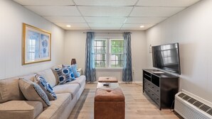 A living room with a sofa, ottoman, TV, and a window with curtains.