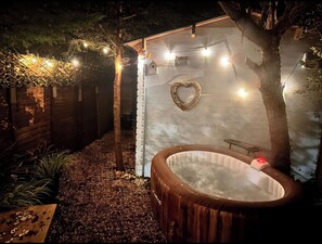 Un jacuzzi en un jardín con valla de madera, luces colgantes y una decoración con forma de corazón en la pared.