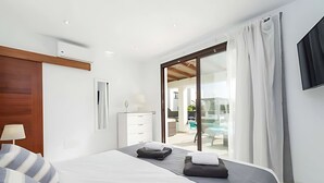 A modern hotel room with a large bed, a wardrobe, a desk, and a view of a pool and outdoor area. - Luxury Villa with Private Pool and Stunning Views in Playa Blanca, Lanzarote (Playa Blanca)