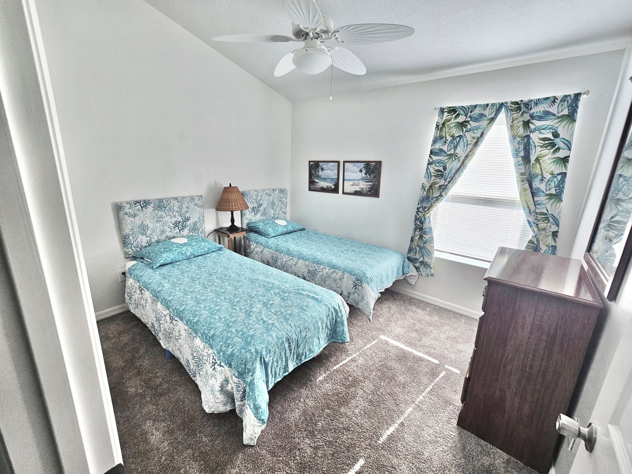 Une chambre avec deux lits, un ventilateur de plafond, une table de chevet avec une lampe, une petite commode et une fenêtre avec des rideaux.