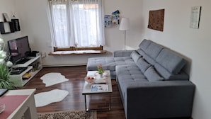 Ett modernt vardagsrum med en grå hörnsoffa, en platt-TV på ett vitt TV-bord, en kristallkrona och ett stort fönster med tunna, genomskinliga gardiner.