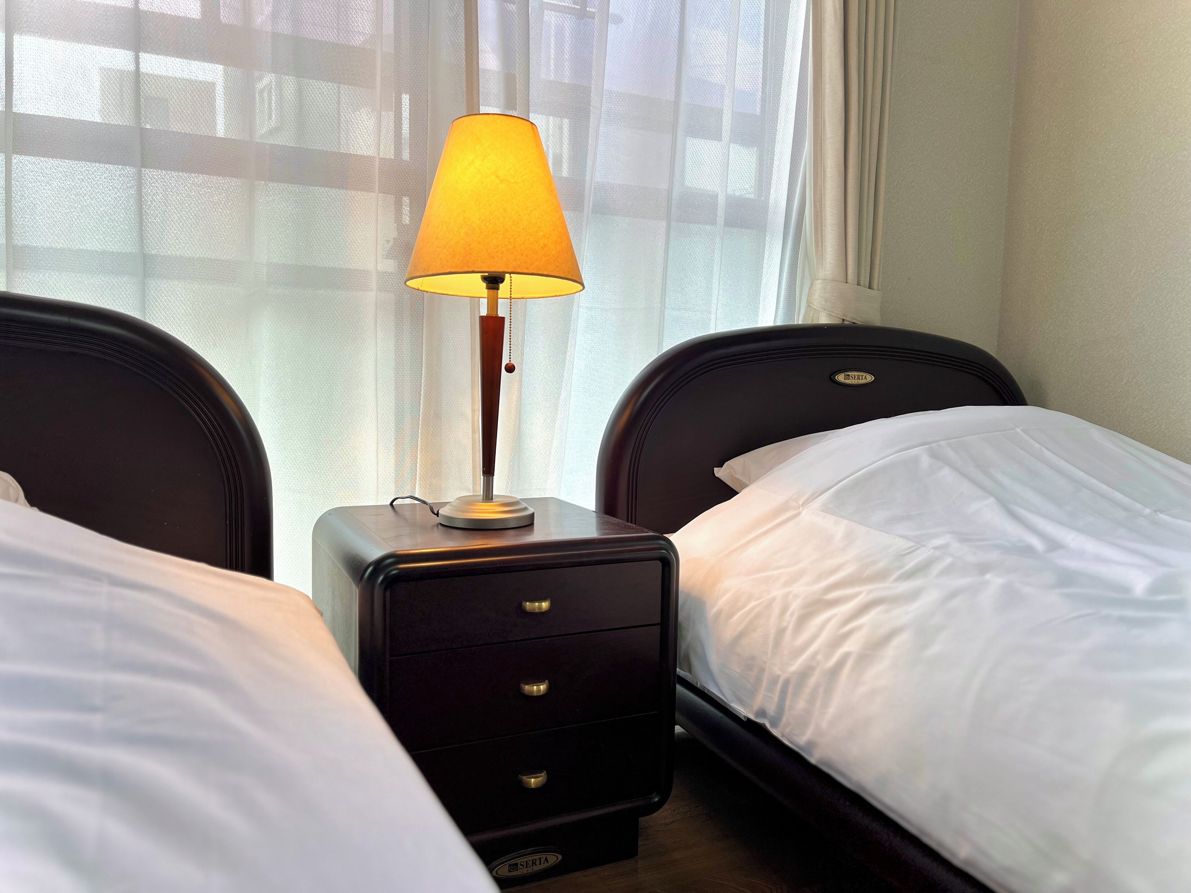 Et hotelværelse med to senge, et natbord med en lampe og lette gardiner.