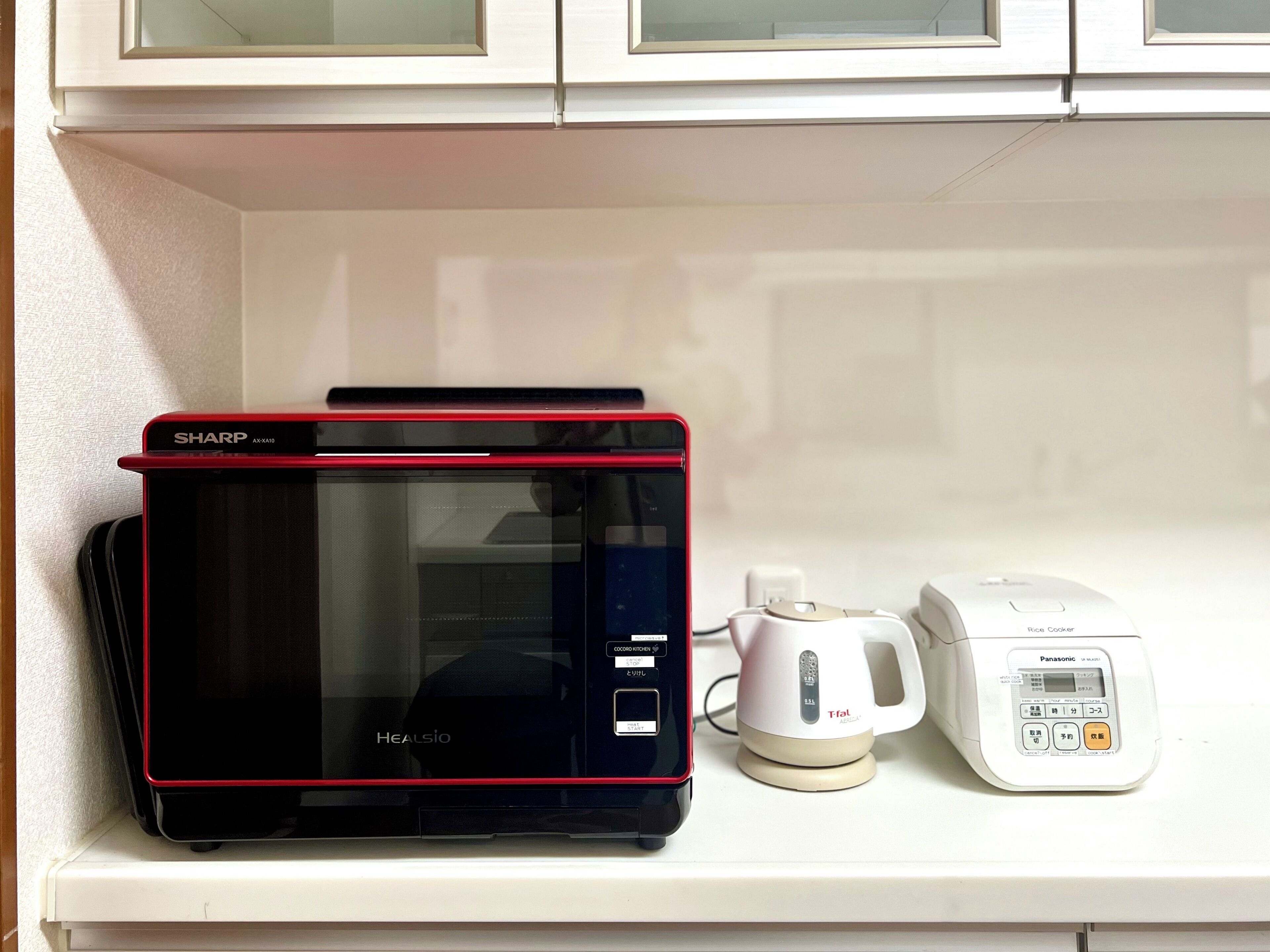 A microwave oven with a red trim, a rice cooker, and a coffee maker on a white shelf.