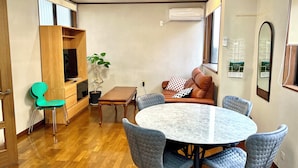 A compact living space with a round dining table surrounded by six chairs, a sofa with cushions, a wooden cabinet, a potted plant, and an air conditioning unit mounted on the wall. - SUMITSUGU HOUSE Station (Kumamoto)