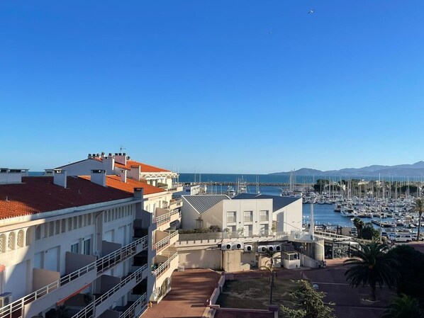 A coastal marina with multiple boats docked, a clear blue sky, and buildings with red-tiled roofs.