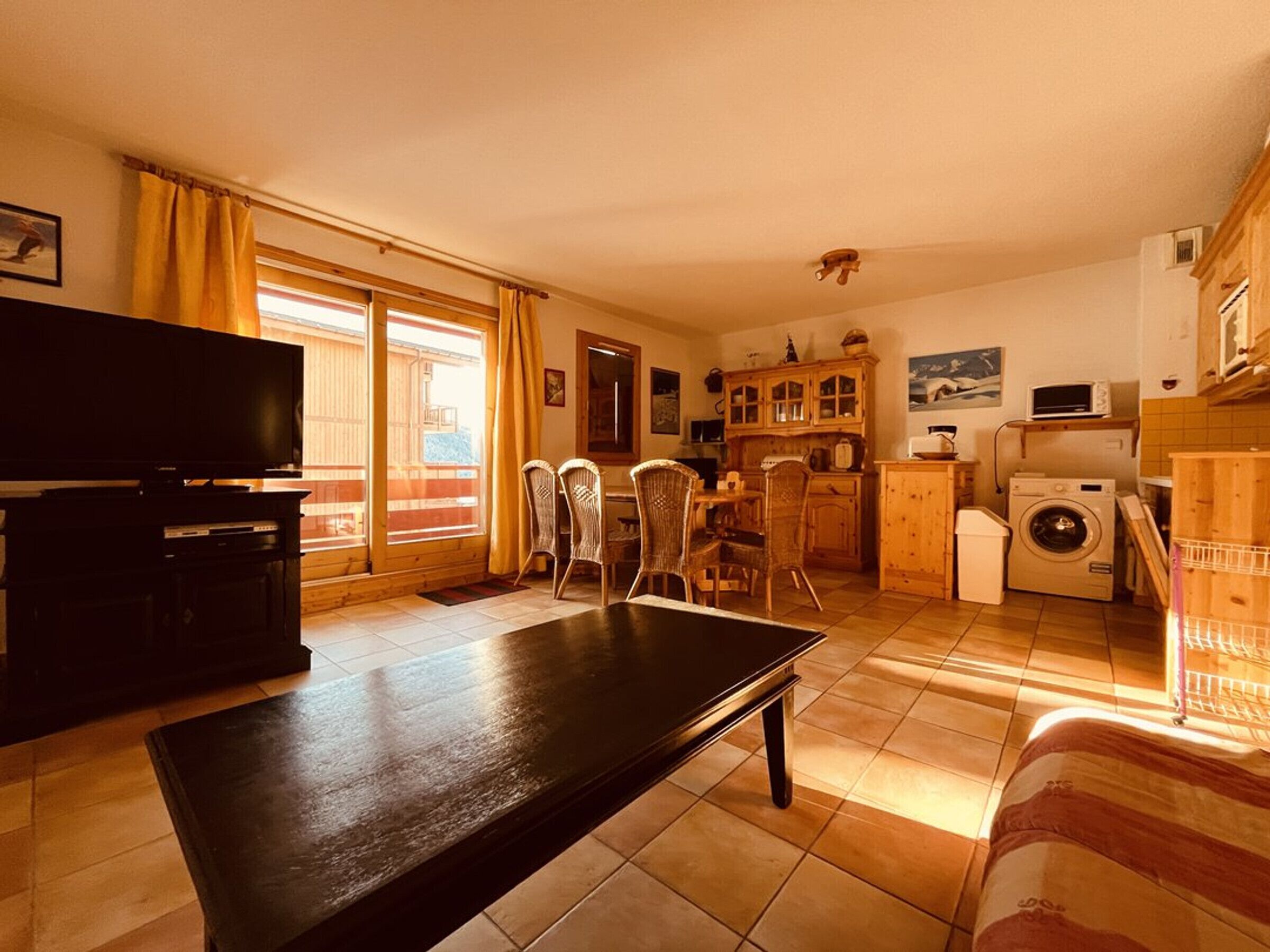 A living room with a TV, a dining area with chairs, a kitchen with a washing machine, and a balcony with yellow curtains.