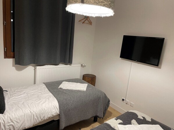 A small, clean room with a bed, a bedside table, a TV mounted on the wall, and a window with curtains.