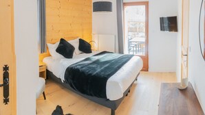 A modern bedroom with a wooden headboard, a bed with white linens and black pillows, a grey blanket, a bedside table, a hanging light, a window with curtains, and a flat-screen TV on the wall. - Beautiful T3 apartment in new residence with parking and WIFI, near village and ski bus (Samoëns)