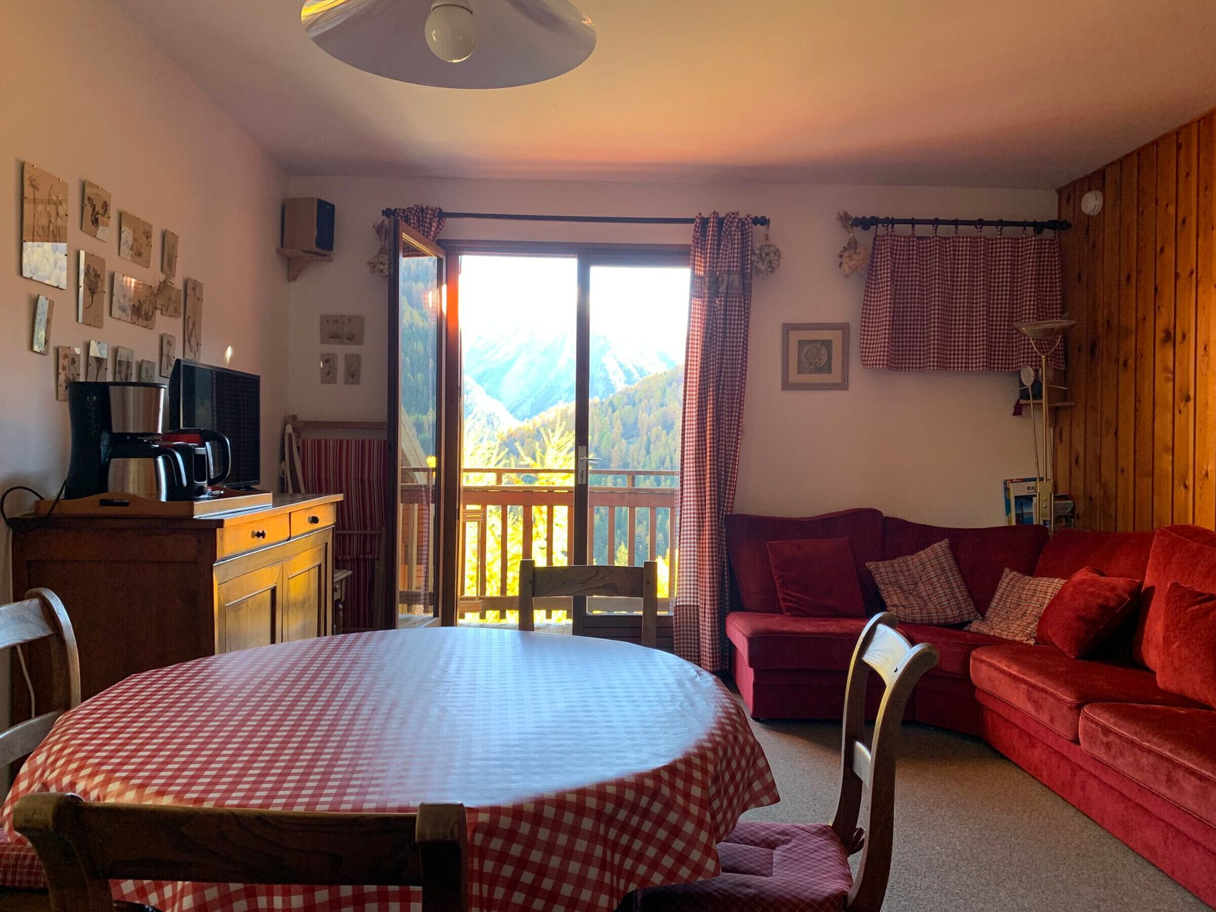 A cozy living room with a red sofa, a red and white checkered table, and a view of mountains through a large sliding glass door.