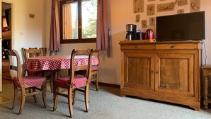 Un coin repas avec une table dressée pour quatre personnes, des chaises en bois, un meuble en bois sur lequel est posée une télévision, et des reproductions botaniques encadrées accrochées au mur.