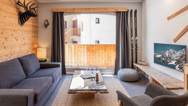 A modern living room with a grey sofa, a wooden coffee table, and a grey armchair. There is a wooden wall panel and a ski rack with skis. - Central apartment with sunny terrace (Le Monêtier-les-Bains)