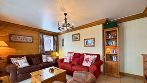 A cozy living room with two sofas, a wooden coffee table, and a red armchair. There is a bookshelf filled with books and a chandelier hanging from the ceiling. - Comfortable apt for 6 in Saint-Martin near slopes, parking, WiFi (Saint-Martin-de-Belleville)
