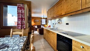 A cozy kitchen with wooden cabinets, a dining area with a patterned tablecloth, and a view of the outside through a window with red curtains. - Comfortable apt for 6 in Saint-Martin near slopes, parking, WiFi (Saint-Martin-de-Belleville)