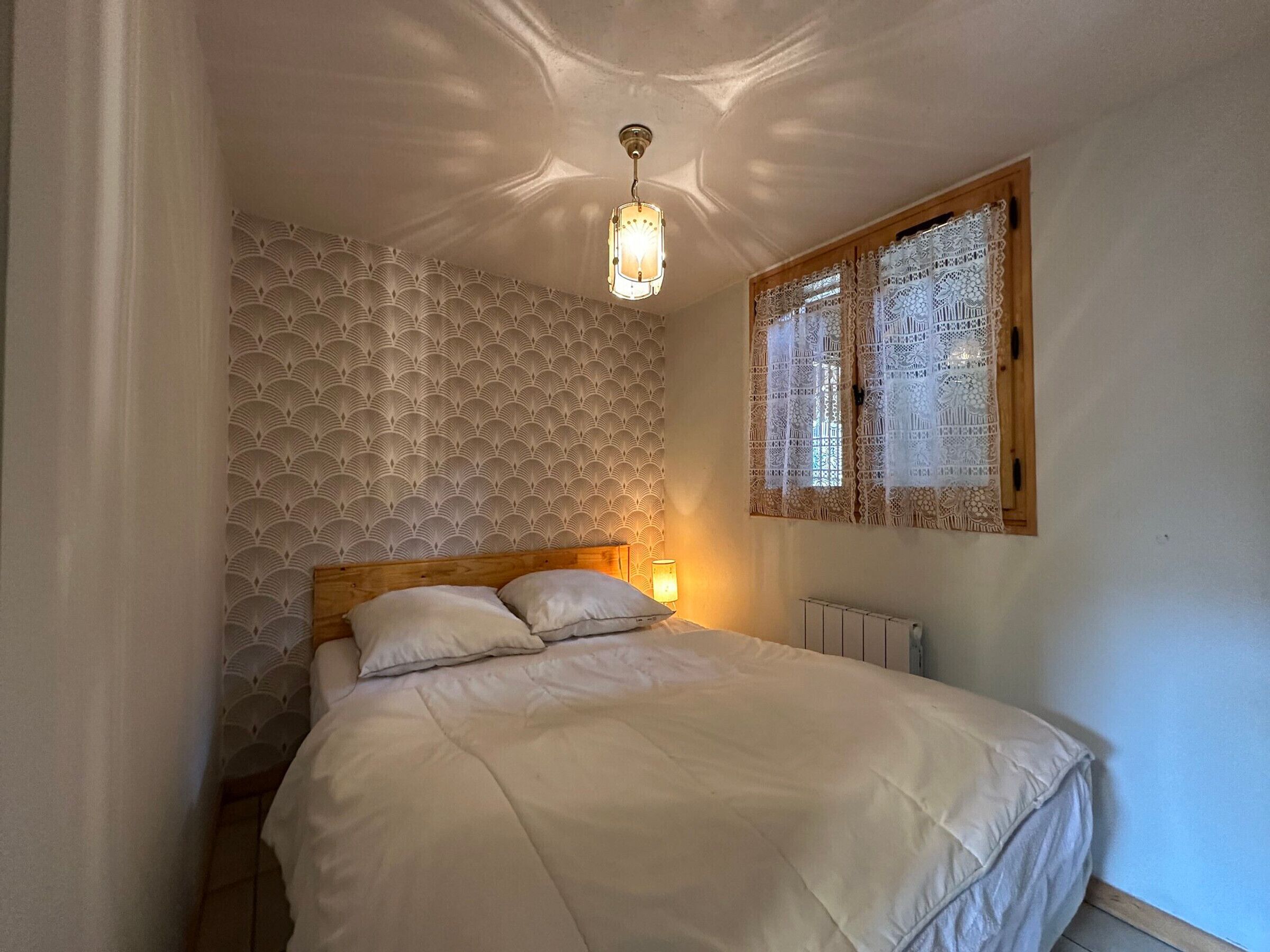 Une chambre à coucher avec un lit, un papier peint à motifs, une tête de lit en bois, une fenêtre avec des rideaux en dentelle et un luminaire suspendu.
