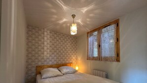 Et soveværelse med en seng, tapet med et mønster, et træhovedgærde, et vindue med blondegardiner og en hængende lampe.