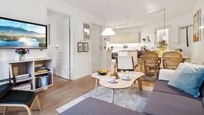 Un appartement moderne comprenant un salon, un coin repas et une kitchenette. On y trouve un téléviseur à écran plat fixé au mur, une bibliothèque et un canapé avec une table basse.