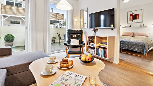 Un salon moderne comprenant un canapé, une table à manger avec des pâtisseries et un magazine, un fauteuil en cuir noir, une bibliothèque, un téléviseur à écran plat et un lit avec une couette grise.