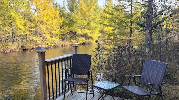 Una terraza de madera con dos sillones de mimbre y una pequeña mesa de vidrio, con vista a un río rodeado de follaje otoñal.