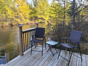 Ein Holzdeck mit zwei Korbstühlen und einem kleinen Glastisch, mit Blick auf einen Fluss, dessen Ufer von Herbstlaub gesäumt sind.